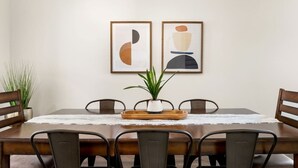 Un comedor con una mesa de madera, seis sillas, dos cuadros abstractos enmarcados, una planta en maceta y un camino de mesa blanco.