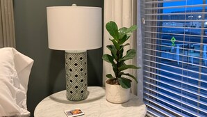 Ein Zimmer mit einem runden Holztisch, einer weißen Lampe mit gemustertem Fuß, einer Topfpflanze und Blick durch Jalousien.
