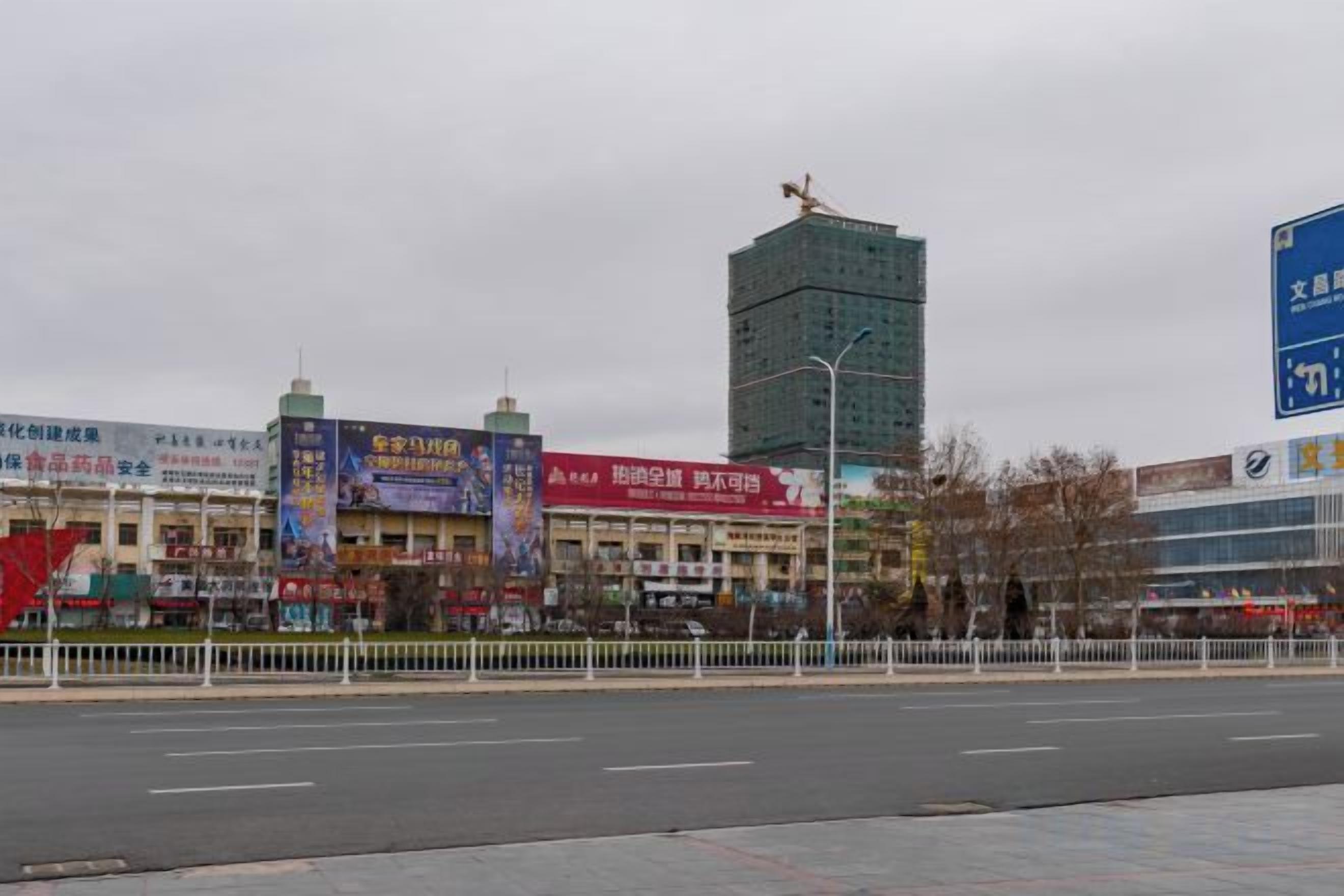 A street view with a tall building, a fence, and various storefronts.