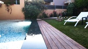 A swimming pool with a wooden deck, surrounded by a grassy area and various plants. - Vista (Saronikos)