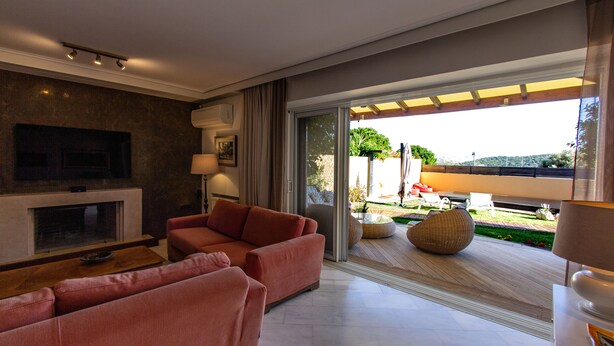 A modern living room with a fireplace, sofas, and a large sliding glass door leading to an outdoor area. - Vista (Saronikos)