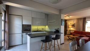 A modern kitchen with a central island, bar stools, and a dining area with a red sofa and wooden table. - Vista (Saronikos)