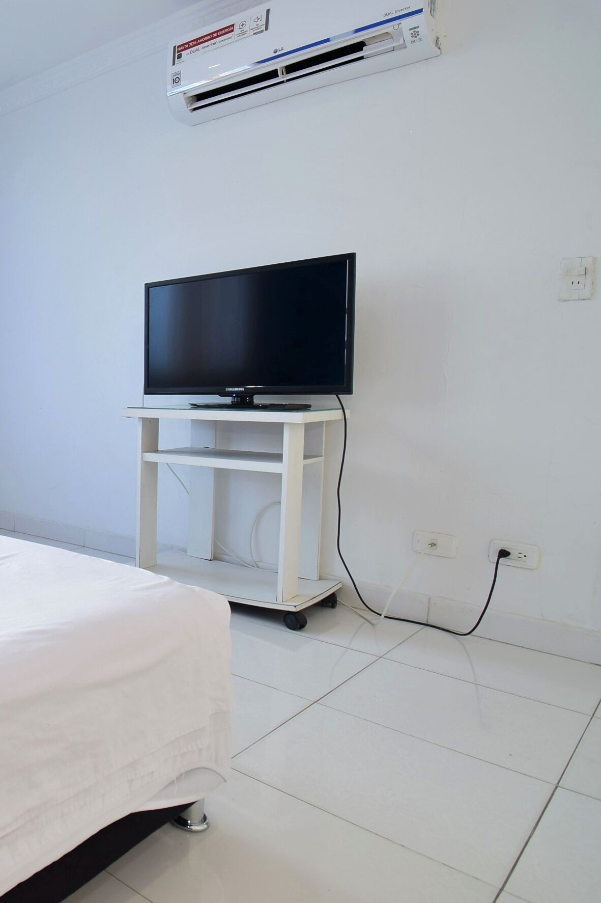 A hotel room with a bed, a TV on a white stand, an air conditioner, and a power outlet.