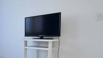 A hotel room with a bed, a TV on a white stand, an air conditioner, and a power outlet.