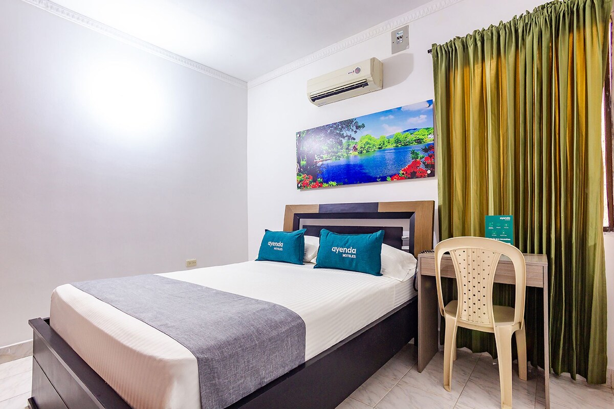 A hotel room with a bed, a desk, a chair, an air conditioner, and a painting on the wall.