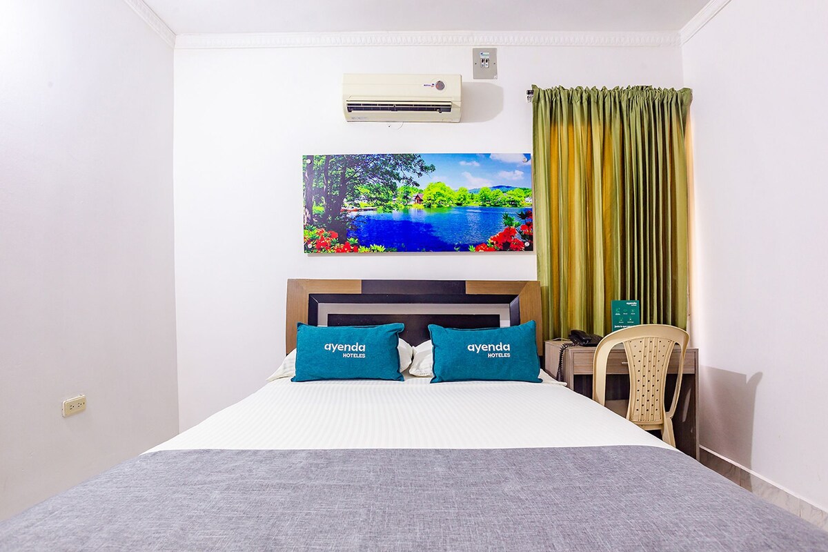 Quarto de hotel com uma cama, ar condicionado fixado na parede, um quadro de um lago e uma cadeira.