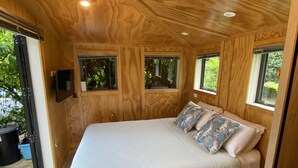 A bedroom with a large bed, wooden walls, and windows with views of greenery. - Tiny Studio - Near New (Whangamatā)