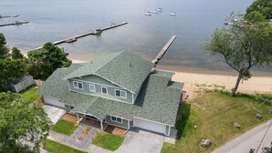 A two-story house with a green roof, a garage, and a driveway leading to a sandy beach. - The Eastside ~ Modern Coastal Getaway w/ Pool, Private Beach, Sauna and Dock (Colchester)