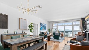 A modern living room with a dining area, large windows, and a view of the city. - The Westside - Hot tub, Private Beach & Dock. Pet-friendly! (Colchester)