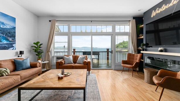 A modern living room with a wooden coffee table, two sofas, a TV, and a balcony with a view of the mountains. - The Westside - Hot tub, Private Beach & Dock. Pet-friendly! (Colchester)