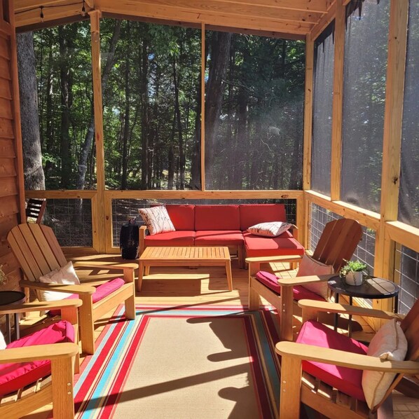 A cozy wooden cabin with a screened porch, red upholstered seating, and a view of a dense forest.