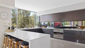 A modern kitchen with a central island, bar stools, a clock, and a view of trees through large windows. - Grandview - Luxurious Entertainer with Spectacular Views (Nelson Bay)