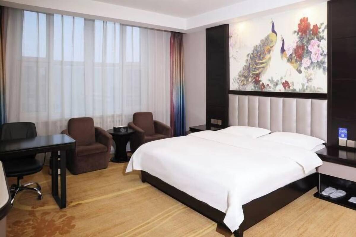 Un cuarto de hotel con una cama grande, un escritorio con silla, una mesita y un cuadro en la pared.