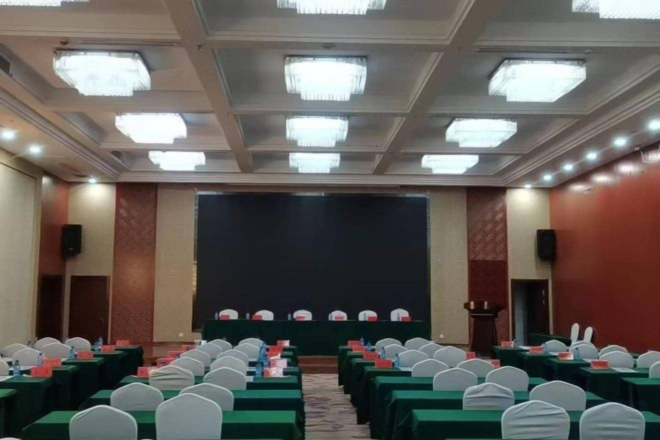 A conference room with rows of tables and chairs, a stage with a podium, and a large screen.