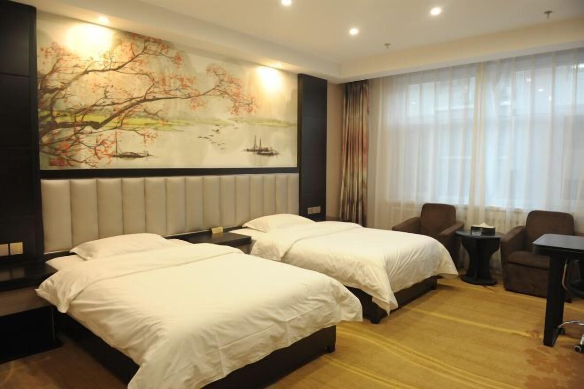 A hotel room with two beds, a large painting on the wall, a view of the outdoors through sheer curtains, and a small table with a chair.