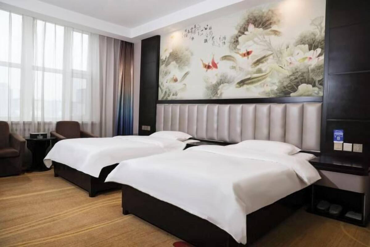 Habitación de hotel con dos camas, un mural grande en la pared, una zona de estar con sillas y una ventana con cortinas.