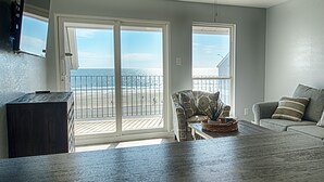 A spacious living room with a large sliding glass door leading to a balcony overlooking a beach.