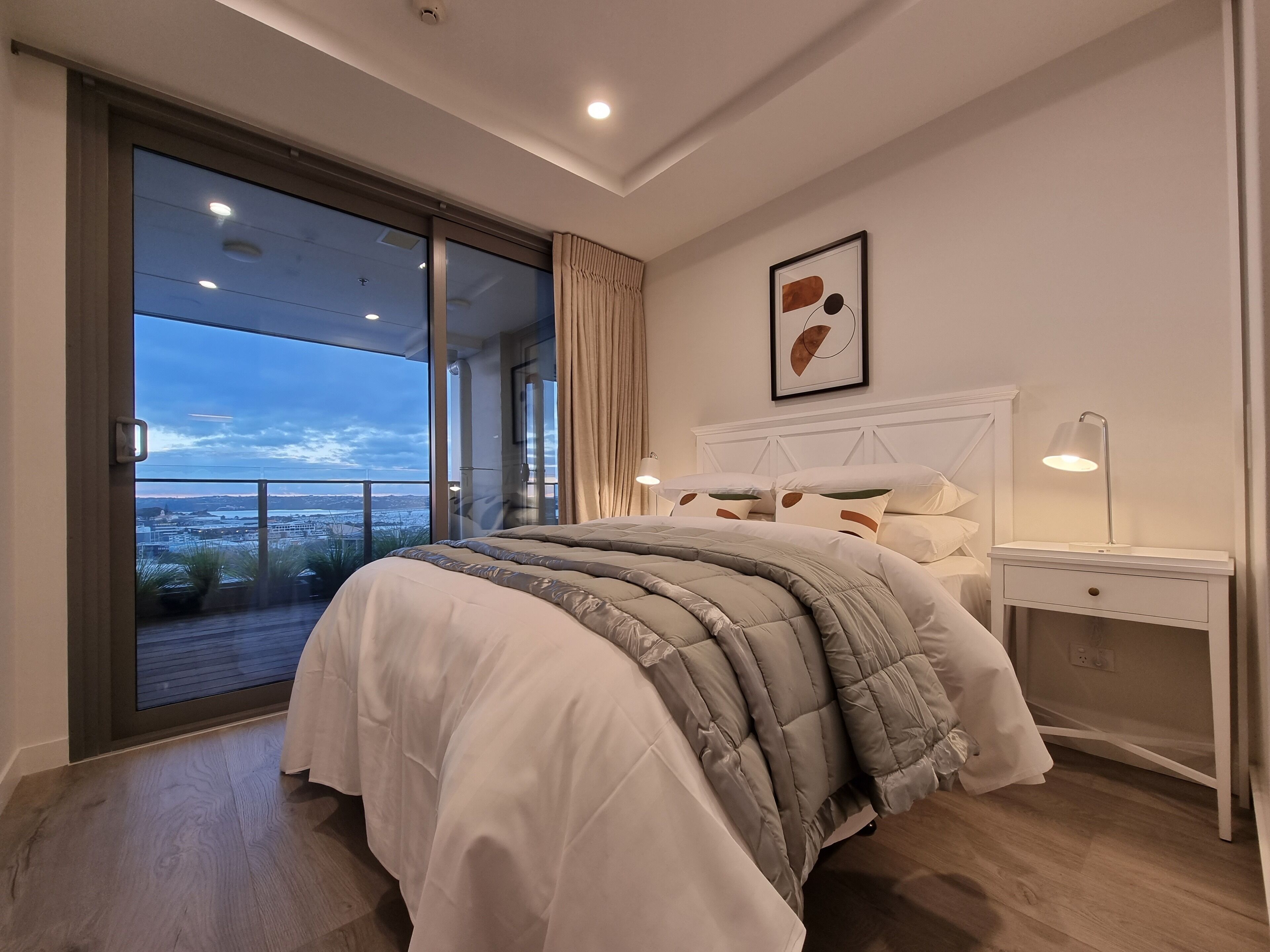 A bedroom with a large bed, a nightstand, a lamp, and a view of the outdoors.