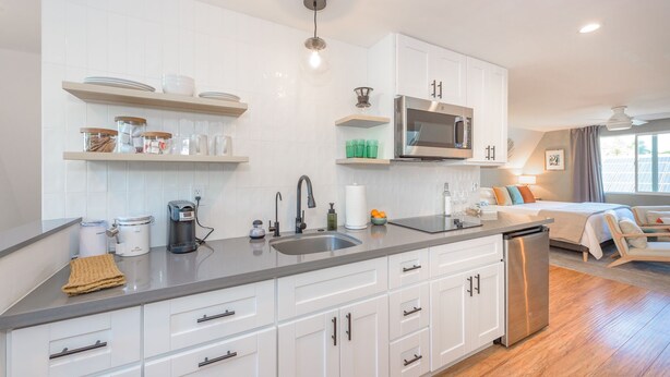 A modern kitchen with white cabinets, a stainless steel refrigerator, and a microwave. There is a sink with a faucet, and shelves above the counter holding dishes and jars.