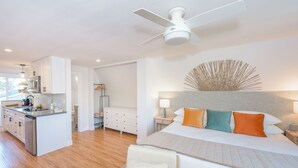 A modern hotel room with a kitchenette, a large bed with multiple pillows, a ceiling fan, and a decorative wall piece.