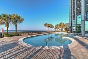 A swimming pool with a curved edge, surrounded by a wooden deck, palm trees, and a building with large windows.