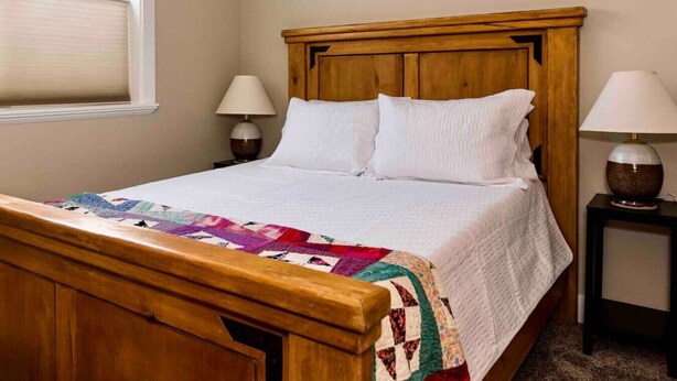A wooden bed with a quilt, two bedside lamps, and a window with blinds. - Montague Manor" W&D, Hot Tub, Queen Beds, Sleeps6 (Bluffdale)