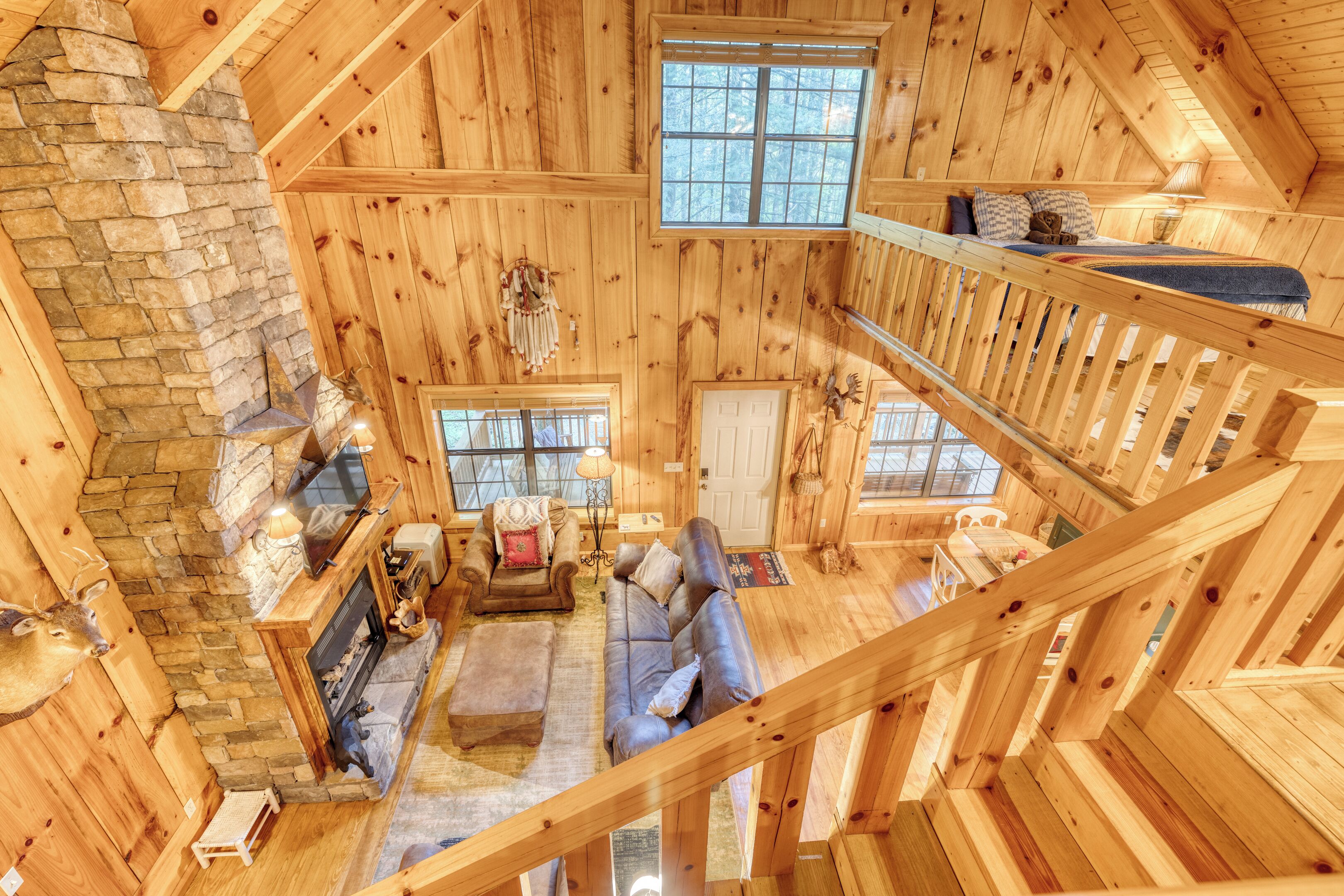 A log cabin interior with a stone fireplace, wooden walls, a loft bedroom with a bed, and a dining area.