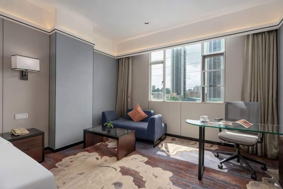 Ein Hotelzimmer mit einem Bett, einem Schlafsofa, einem Glastisch, einem Schreibtisch mit Stuhl und Blick auf die Stadt durch große Fenster.