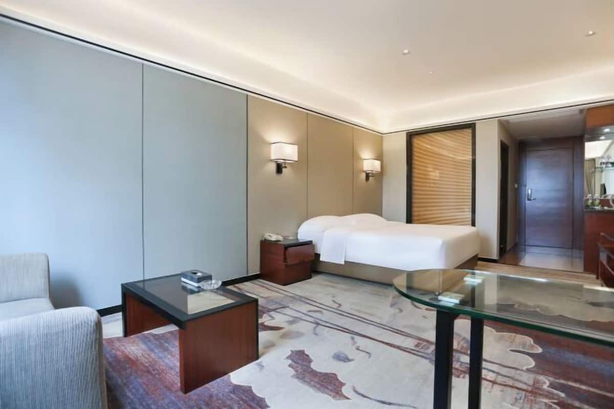 Ein modernes Hotelzimmer mit einem Bett, einem Glastisch, einem Sessel, einem Nachttisch und einer Wandlampe.