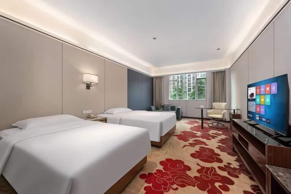 Ein Hotelzimmer mit zwei Betten, einem Fernseher, einem Schreibtisch, einem Sessel und einem Fenster mit Blick.