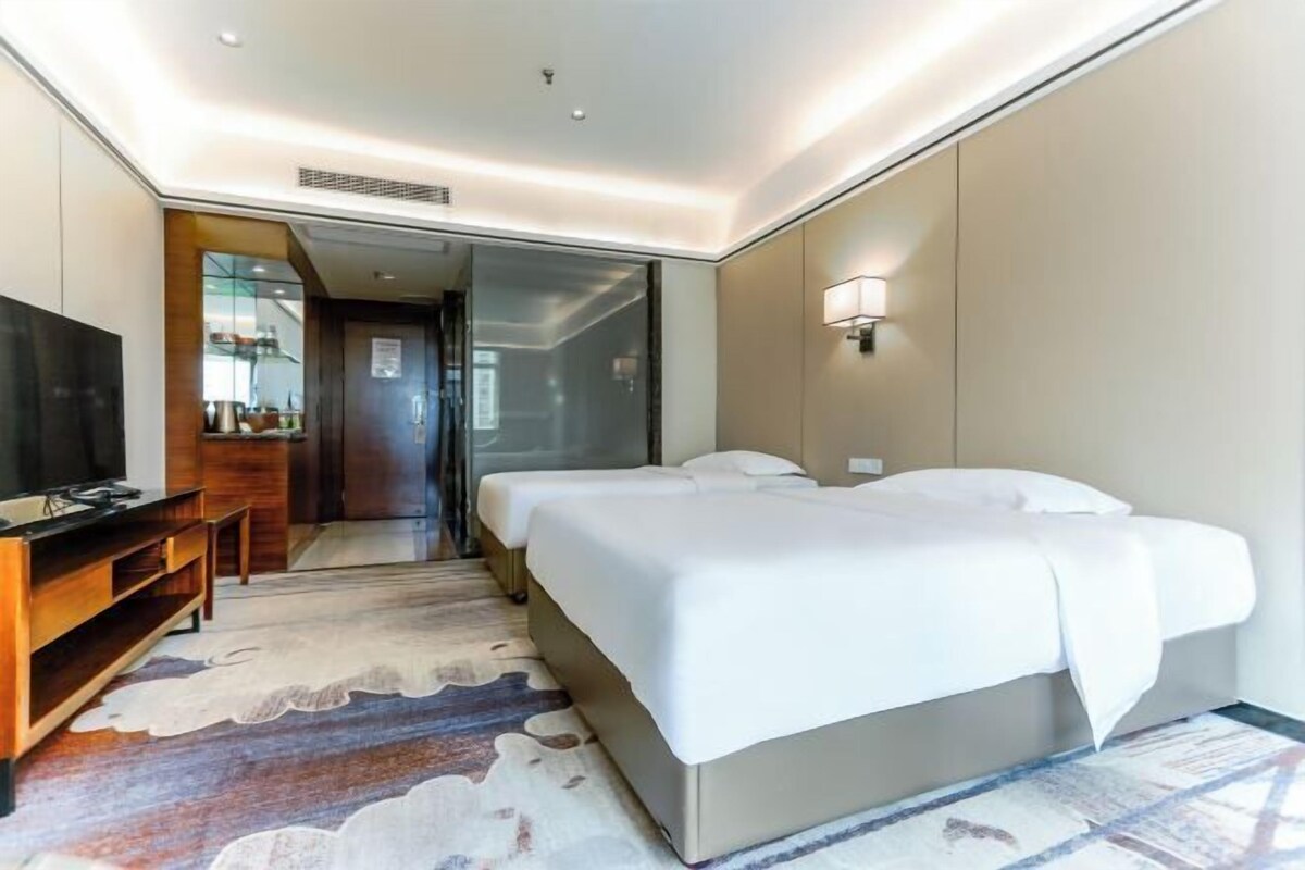 A hotel room with a large bed, a TV, a desk, and a bathroom visible through a glass door.