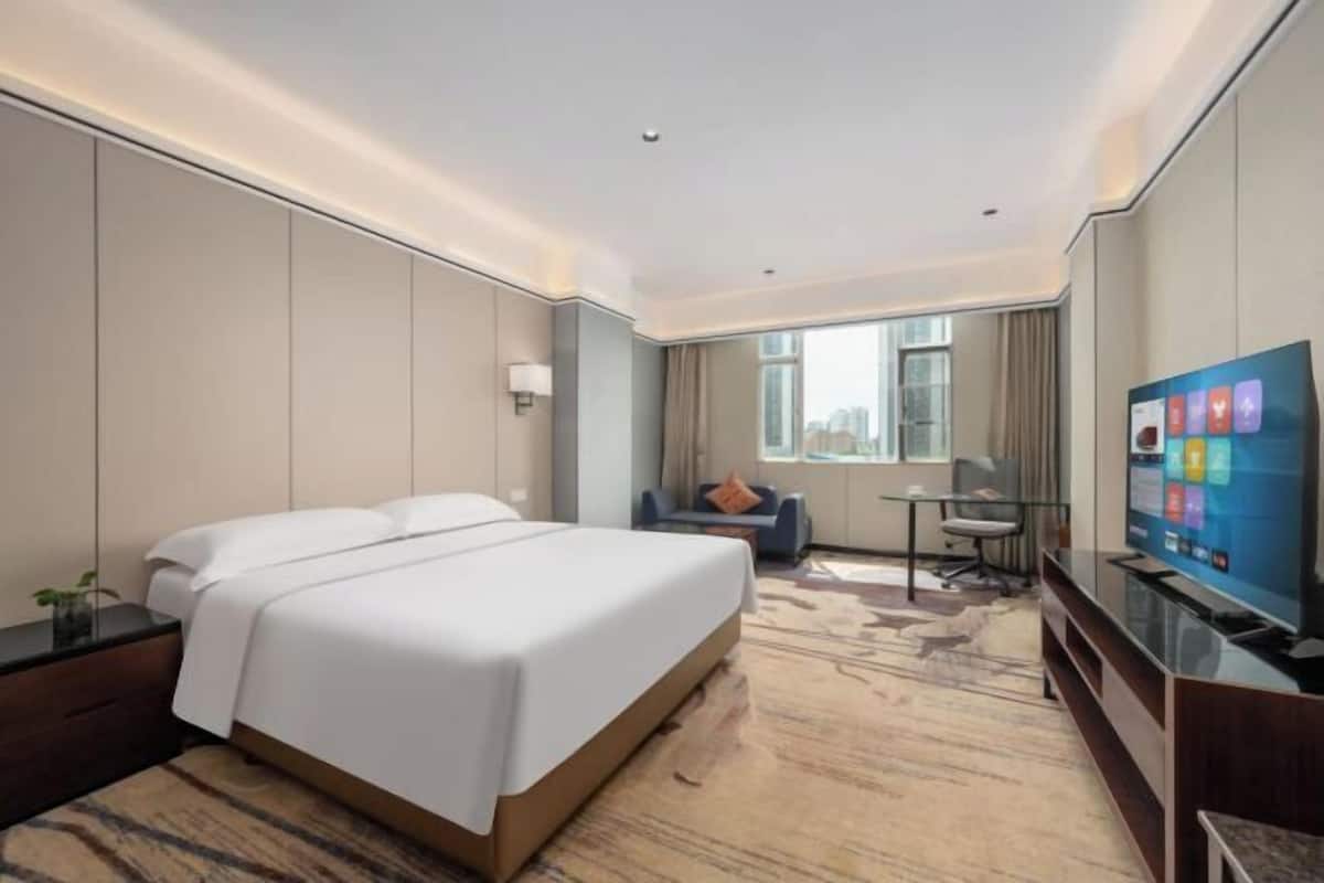 Ein Hotelzimmer mit einem großen Bett, einem Schreibtisch, einem Sessel, einem Fernseher und einem Fenster mit Blick auf die Stadt.
