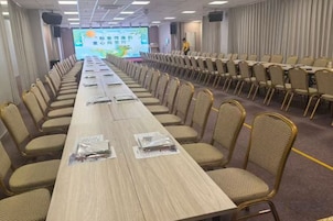 Un salón de conferencias con una larga mesa, filas de sillas y una pantalla de proyección.