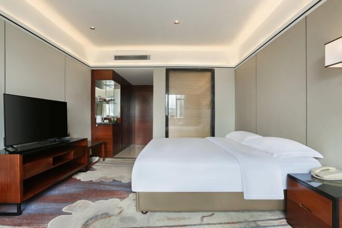 A modern hotel room with a flat-screen TV, a bed with white linens, a wooden nightstand, and a desk with a telephone.