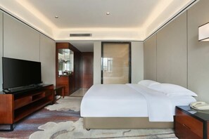 Quarto de hotel moderno com televisão de tela plana, cama com roupa de cama branca, mesa de cabeceira de madeira e escrivaninha com telefone.