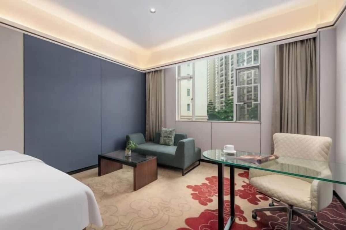 Ein Hotelzimmer mit einem Bett, einer Couch, einem Glastisch, einem Sessel und einem großen Fenster mit Vorhängen.