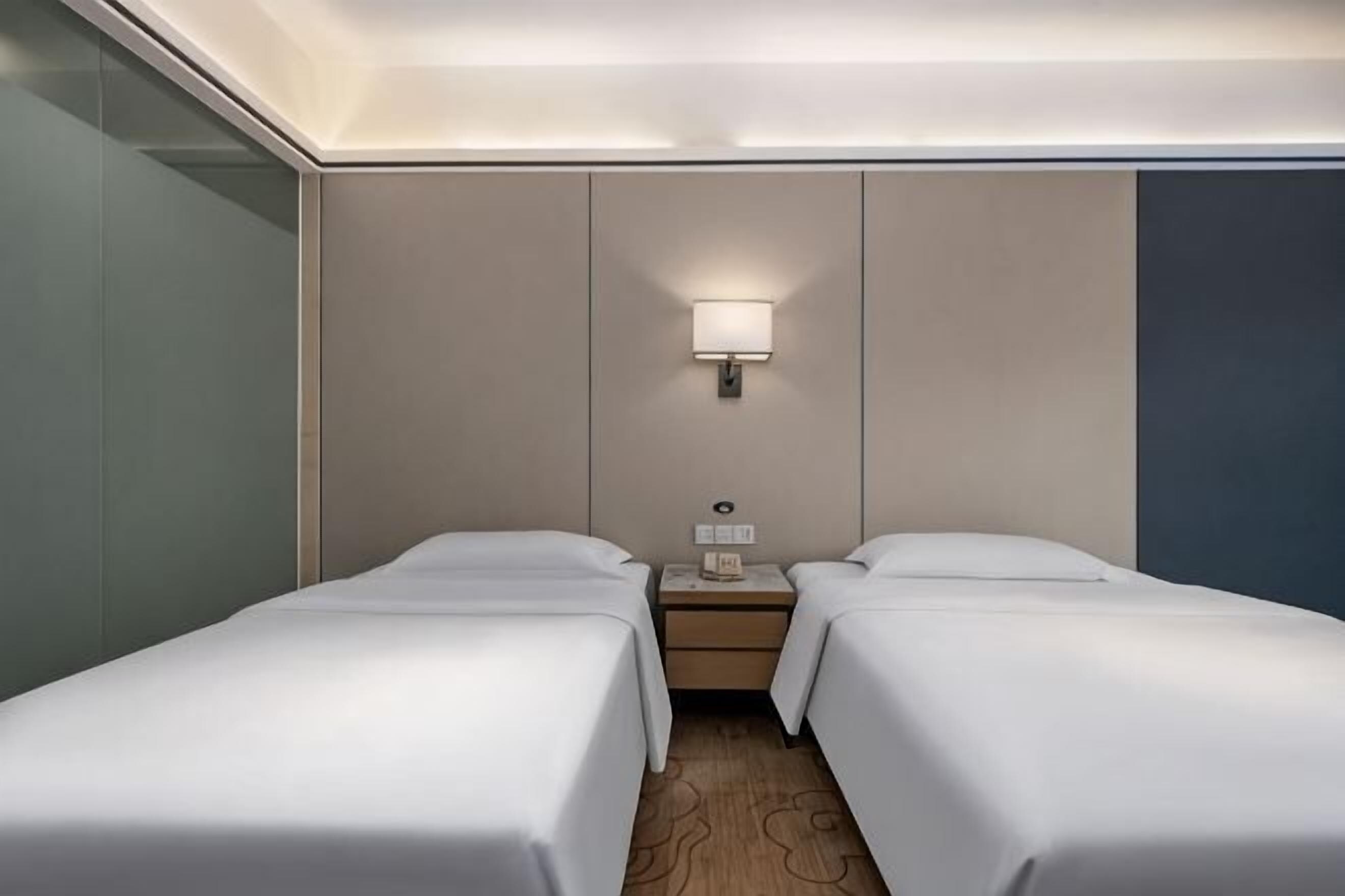 A hotel room with two single beds, a nightstand, a lamp, and a wall-mounted light fixture.