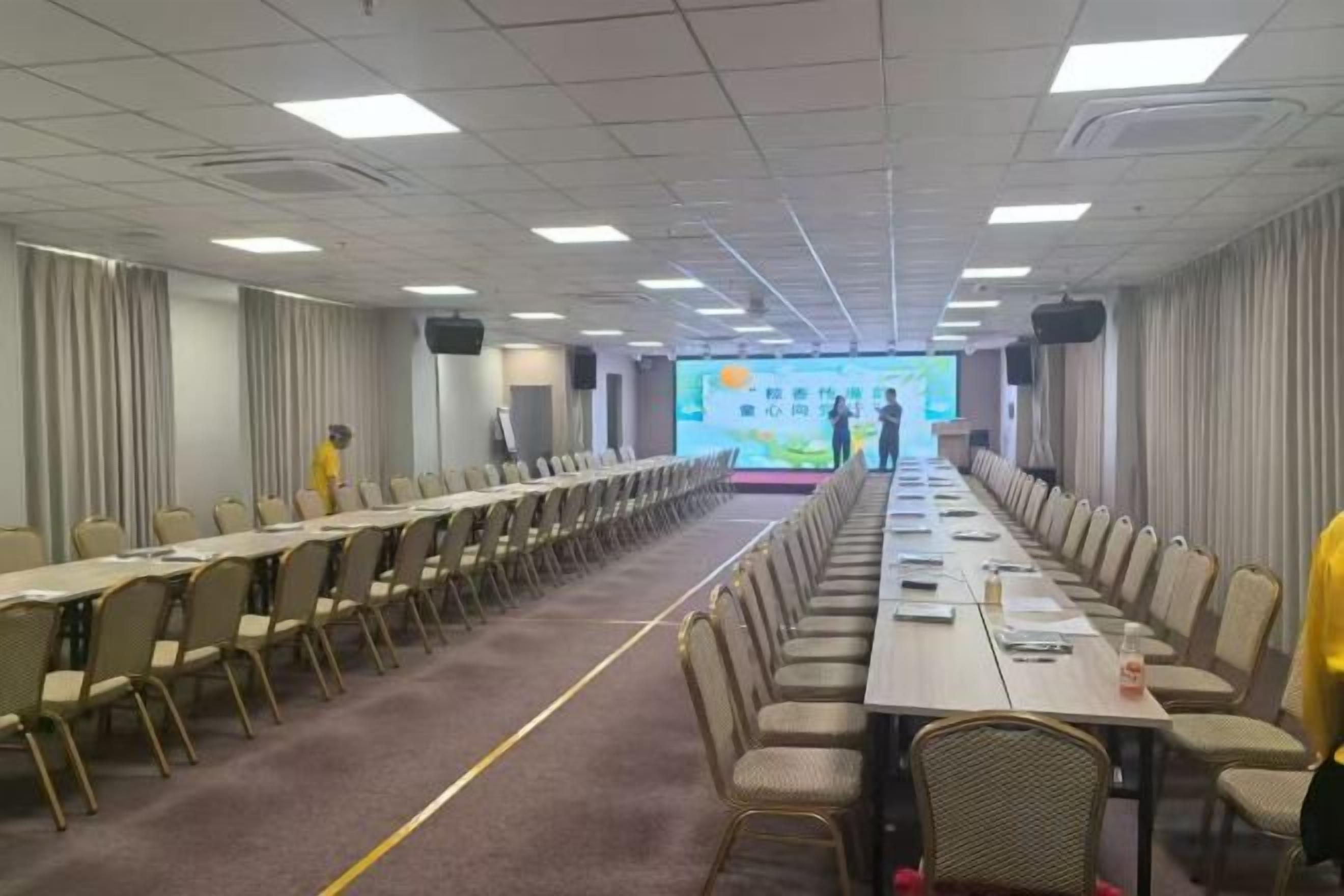 A conference room with long tables and chairs, a stage with a presentation screen, and a carpeted floor.