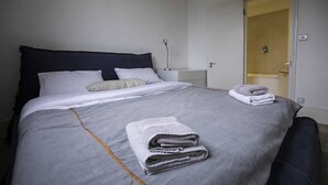 Un llit ben fet amb un estovat i coixins grisos, una tovallola doblada al llit i una porta que condueix a un bany.
