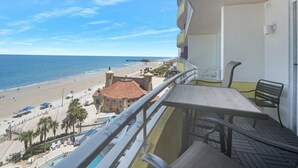 A balcony with a view of the beach, a pool, and a building with a red-tiled roof.