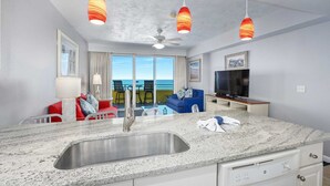 A modern kitchen with a granite countertop, stainless steel sink, and a ceiling fan. There is a dining area with a red couch and a blue sofa. A flat-screen TV is mounted on the wall.
