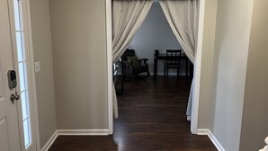 Un pasillo con una puerta a la izquierda, una zona de comedor con mesa y sillas en el centro, y otra puerta blanca a la derecha.