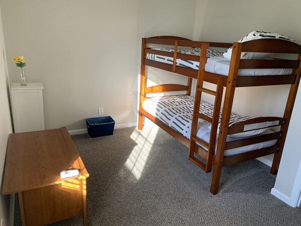 A bunk bed with a patterned bedspread, a small blue storage bin, a wooden nightstand with a lamp, and a vase with yellow flowers on a white cabinet. - Wingate Estates (Spring Hill)