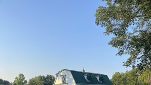 Une grange avec un toit vert et un revêtement blanc, entourée d’une clôture blanche et bordée d’un chemin de gravier.