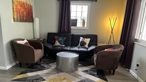 Ein Wohnzimmer mit einem schwarzen Ledersofa, zwei braunen Sesseln, einem grauen, strukturierten Ottomanen und einem gemusterten Teppich.