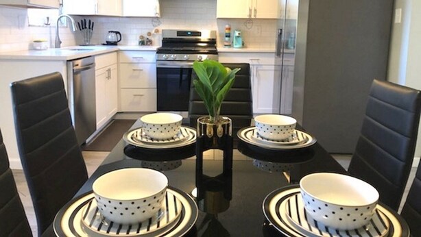 Eine moderne Küche mit weißen Schränken, einem schwarzen Herd und einem Kühlschrank. Ein Esstisch mit vier Stühlen und eine Vase mit Blumen steht bereit.