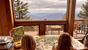 Deux personnes sont penchées sur un puzzle, assis à une table près d’une fenêtre qui offre une vue sur les montagnes.
