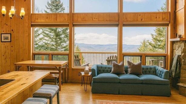 L’intérieur chaleureux d’un chalet avec une grande table à manger en bois, un canapé bleu agrémenté de coussins et une vue sur les montagnes par de grandes fenêtres.