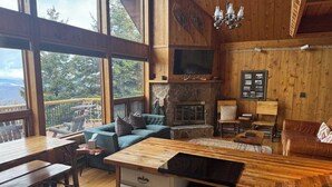 L’intérieur chaleureux d’un chalet avec une cheminée en pierre, des meubles en bois et de grandes fenêtres offrant une vue panoramique.