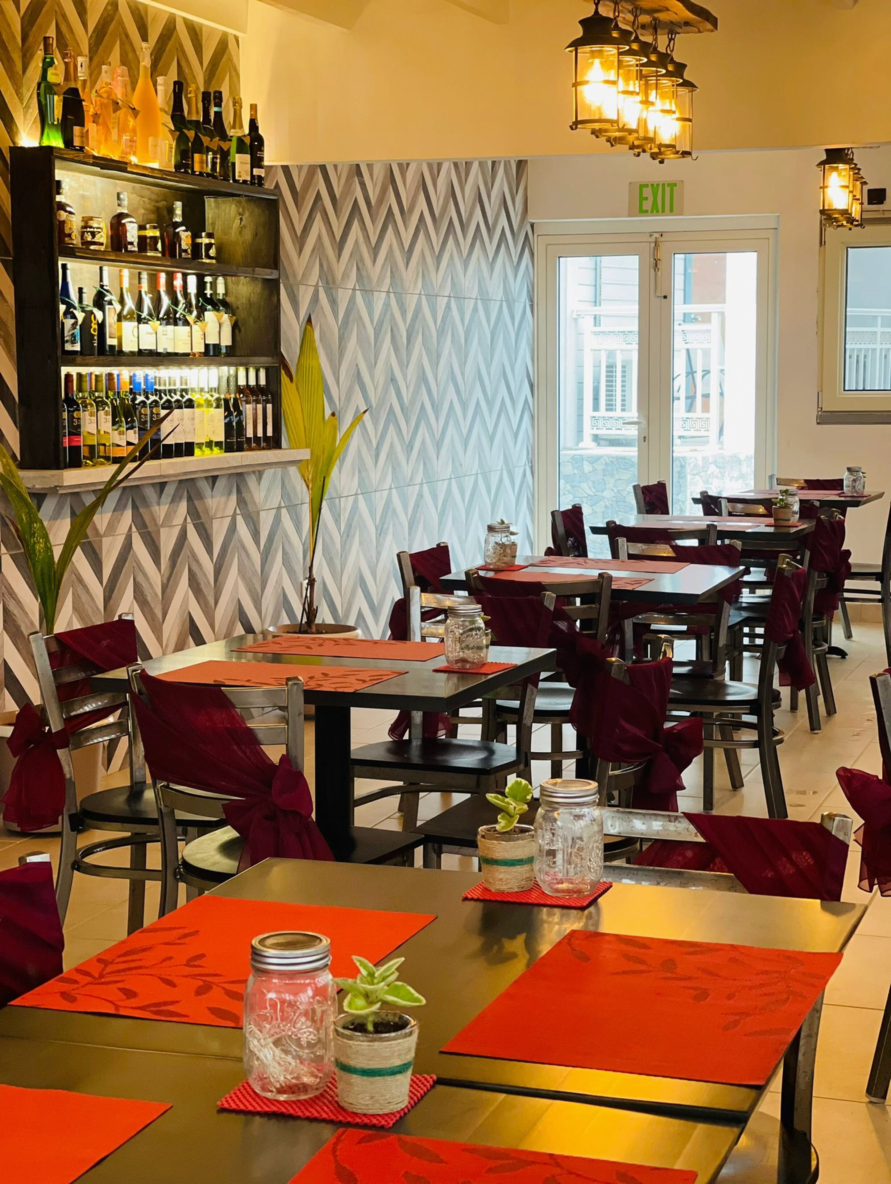 A restaurant with a bar area displaying various bottles, tables with red tablecloths and chairs, and a herringbone patterned wall.